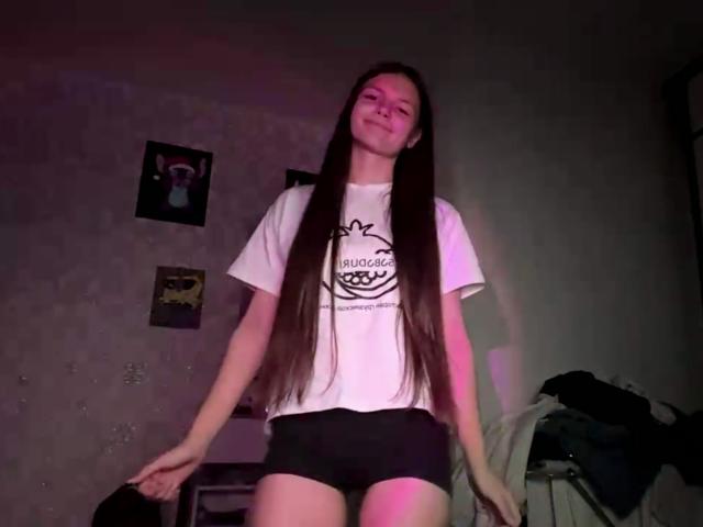 EleonoraMcaleese with long brown hair, white t-shirt, black shorts, standing, smiling, pink and purple lighting, white walls, wall posters, clothes pile on right.