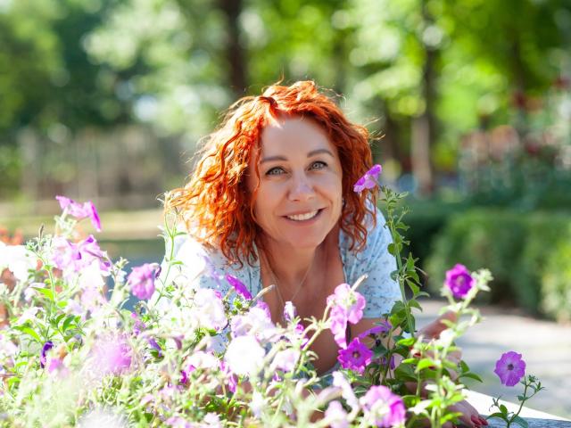TaylorFoxT with curly red hair, smiling, leaning on a bed of purple and pink flowers, green eyes, white shirt, sunlit garden, bright greenery in the background, relaxed.