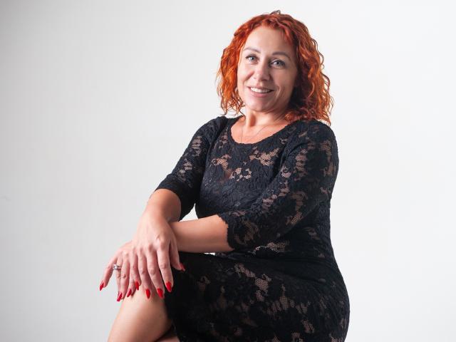 TaylorFoxT with red curly hair, black lace dress, red nails, smiling, sitting, legs crossed, hands on knee, white background, soft light, cute, sexy, medium-sized tits, attractive.