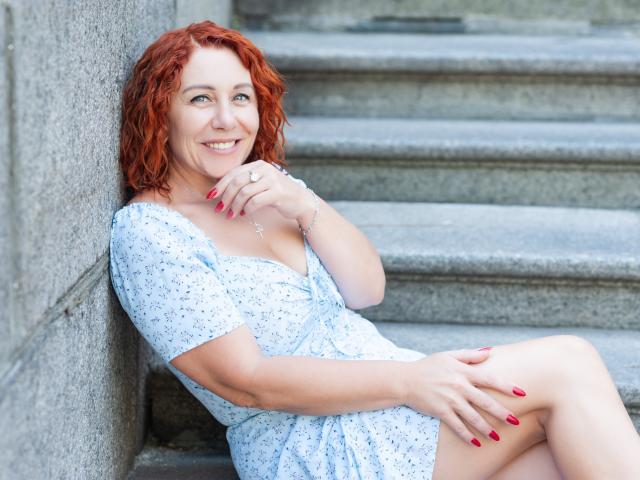 TaylorFoxT with red curly hair, smiling, sitting on stone steps, wearing a white floral dress, red nails, touching chin, showing cleavage, hand on knee, casual and cute.