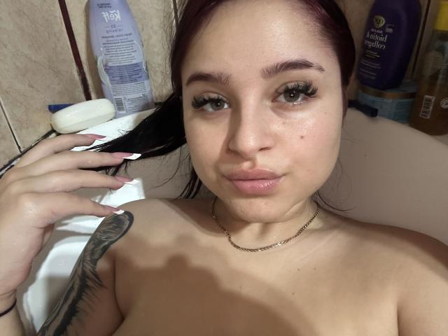 Luucie with red hair, green eyes, and a tattoo on her right shoulder, topless in a bathtub, holding her black hair, wearing a chain necklace, pouting slightly.
