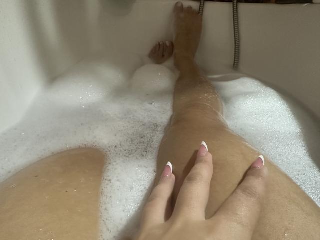 Las piernas de Luucie en una bañera, cubiertas de espuma blanca, con su mano apoyada en su muslo. Tiene uñas rosas con puntas francesas y sus dedos de los pies son visibles por encima de la espuma..