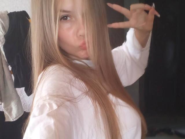 RuthFleetwood with long blonde hair, pouty lips, and a white shirt, making a peace sign with her right hand, showing her tits slightly. Soft, natural light.