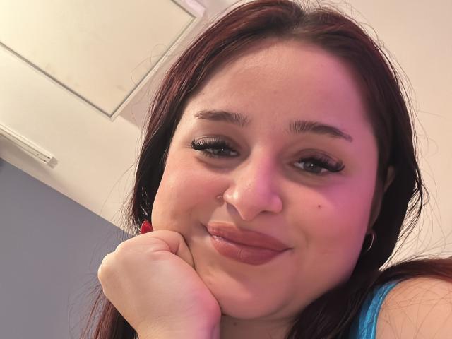 Luucie has dark brown hair, full eyebrows, and is smiling with her hand under her chin, showing off her pink lips and long eyelashes. She's wearing a blue top.