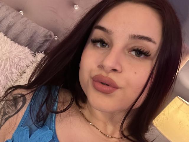 Luucie has puffy pink lips, long brown hair, blue lace top, and a tattoo on her left arm. She's wearing a gold chain necklace, looking directly at the camera.