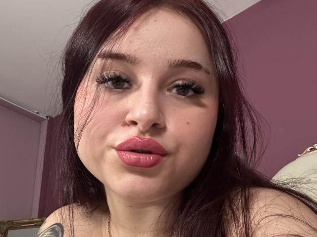 Luucie has puffy pink lips, dark red hair, and long eyelashes. She's pouting slightly with a small tattoo on her shoulder. Her skin is fair with a few moles.