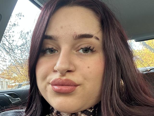 Luucie with pouty pink lips, dark brown hair, green eyes, and a nose piercing, sitting in a car, wearing a black and white patterned scarf, looking at the camera.