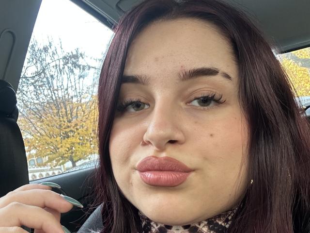 Luucie has puffy pink lips, dark red hair, and freckles. She's making a pouty face with her hand near her cheek, sitting in a car.