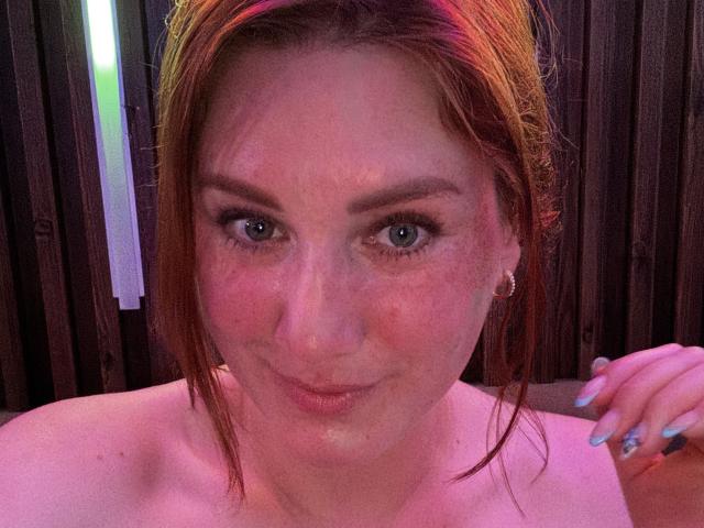 KatyaRogersX has wet red hair, light skin, blue eyes, and a slightly flushed face with visible cum on her cheek and upper chest. She's wearing small earrings and has blue nails.