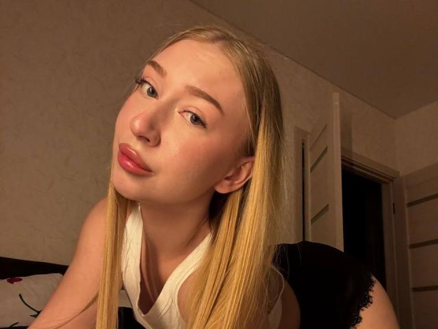 JoetteShigeta with long blonde hair, pouty pink lips, and light skin, wearing a white top, looking at the camera with a slight tilt to her head, in a dimly lit room.