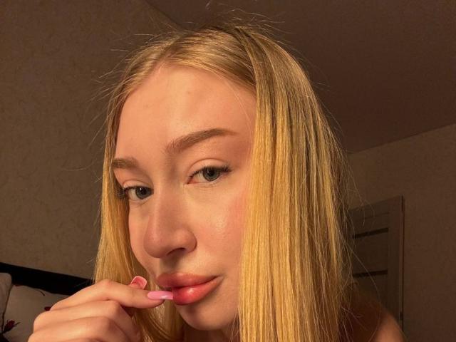 JoetteShigeta with blonde hair, blue eyes, and pink lipstick, biting her pink fingernail, showing a slight smile, in a warm, softly lit room.