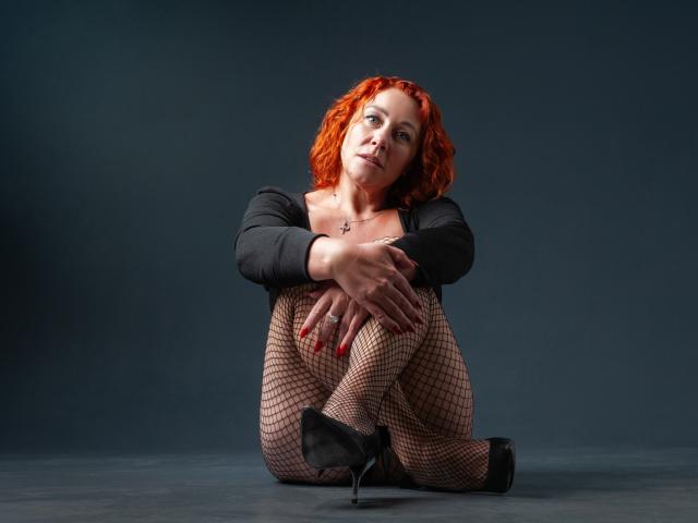 TaylorFoxT with red hair, black top, fishnet pants, and black heels, sitting with knees up, arms around legs, looking at camera, hands on knees, red nails.