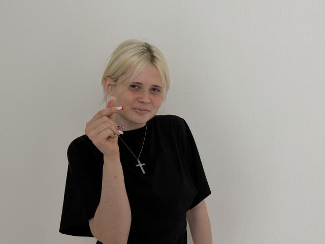 Blond XochitlAlejo with a cross necklace, wearing a black shirt, holding a purple earring, slightly smiling, and raising her left hand.