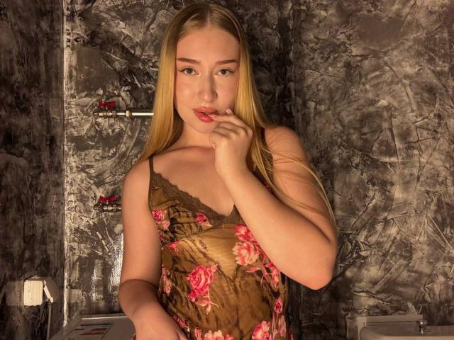 Joette Shigeta, com cabelos loiros longos, vestindo um vestido floral marrom, chupando o dedo, exibindo os seios, em um banheiro com pouca luz e paredes de mármore escuro..
