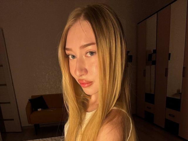 JoetteShigeta with long blonde hair, blue eyes, and red lips, looking at the camera, wearing a white top, in a warmly lit room. Boobs not visible.