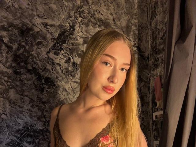 Die blonde JoetteShigeta mit langen Haaren, die ein braunes Spitzenoberteil trägt, hat kleine Brüste, helle Haut und rosafarbene Lippen und blickt leicht zur Seite mit einem leichten Lächeln..