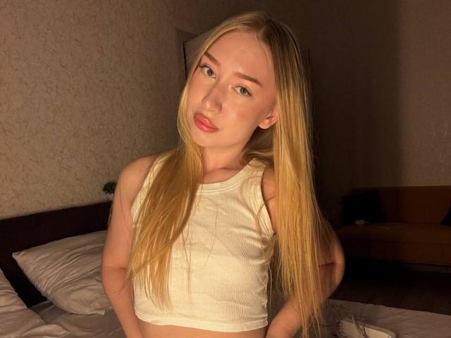Blonde JoetteShigeta with long hair, wearing a white crop top, standing in a dimly lit room, hands on hips, looking at camera with slightly parted pink lips.