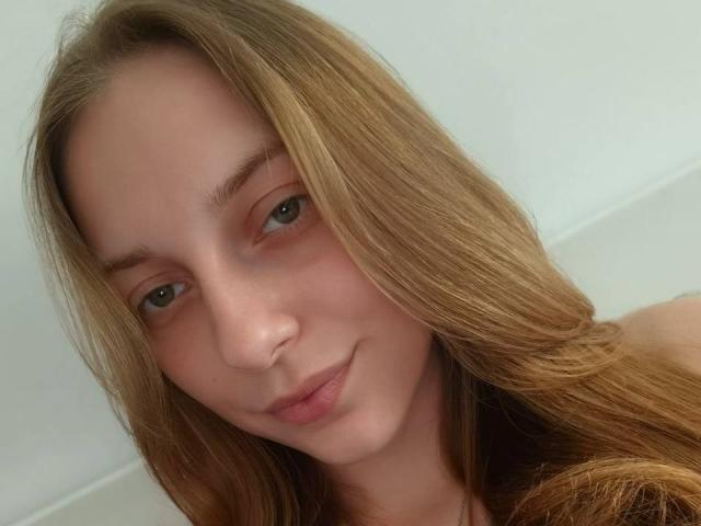 Aizza has light brown, straight hair, green eyes, and a slight smile, with fair skin and no makeup. She's looking at the camera with a relaxed expression.