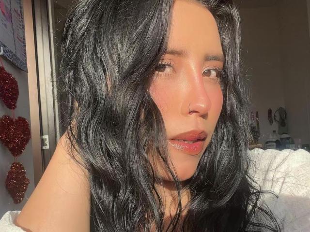 Elizabeth Macy ha i capelli neri e mossi, gli occhi marroni e le labbra rosa lucide. Guarda leggermente di lato, con la luce del sole sul viso, e indossa un piccolo piercing al naso..
