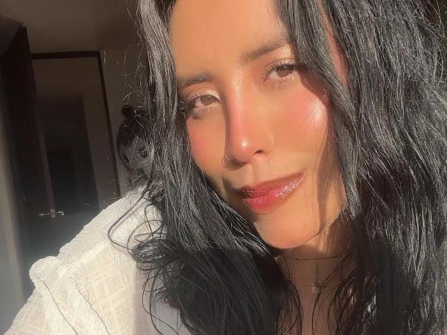 ElizabethMacy has shiny, sunlit black hair, pinkish cheeks, and glossy red lips. She's wearing a white shirt, with a soft, confident smile. Her face is slightly angled.