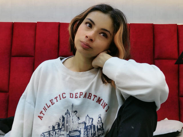 Amuneet, con cabello castaño, labios carnosos, vistiendo una sudadera blanca del departamento de deportes, con la cabeza apoyada en la mano, sentada en una cama con cabecera tapizada de color rojo, mirando hacia un lado..