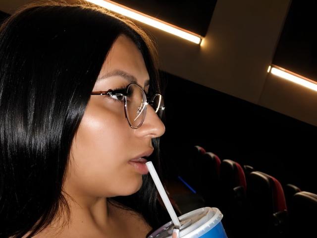 Catalina Rivero mit schwarzen Haaren, Brille und einem Getränk mit Strohhalm, die daran nippt, ihr Gesicht leuchtet im schwachen Licht, ihr Hintern ist kaum im dunklen Hintergrund zu erkennen..