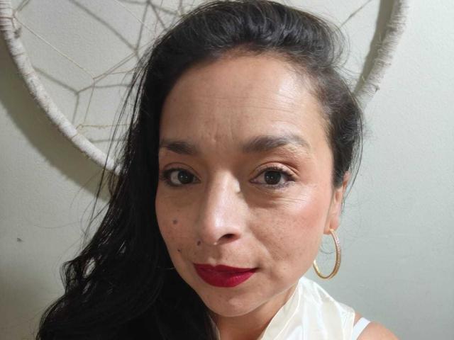 StangelarRivera with dark hair, red lipstick, small mole on left cheek, and gold hoop earrings, wearing a white top, has a neutral expression, and a dreamcatcher behind her.