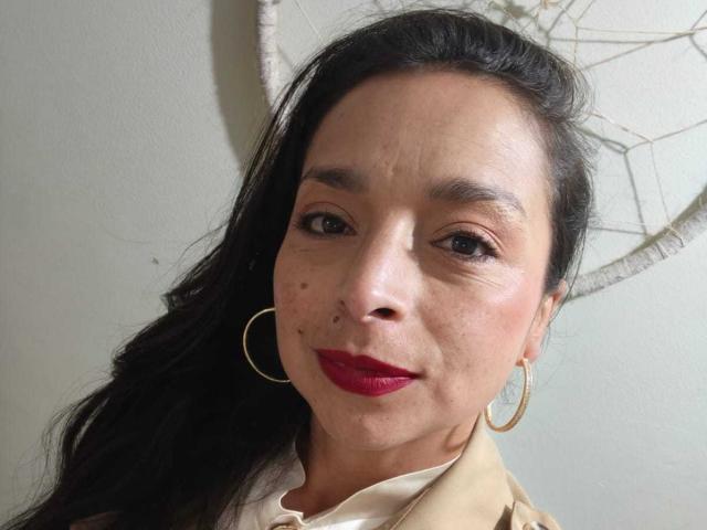 StangelarRivera has medium brown skin, black hair, red lipstick, small moles on her face, and wears gold hoop earrings. She's smiling slightly with a beige shirt.