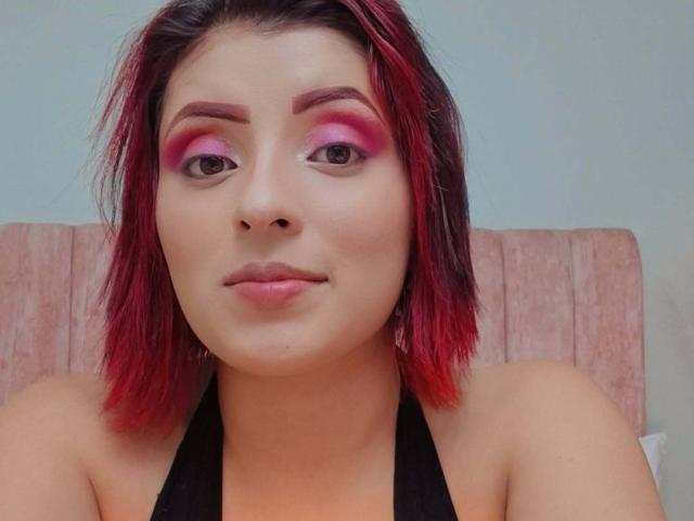LizBonnet with red and black shoulder-length hair, pink eyeshadow, and black halter top, has a neutral expression and fair skin. Her tits are not visible.