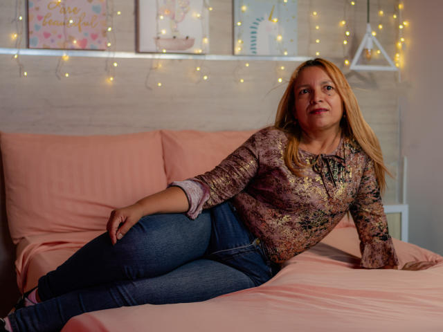 SandyMatture is a middle-aged woman with blonde hair, wearing a floral shirt and blue jeans, lying on a pink bed, her right arm behind her, smiling gently.