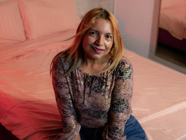 SandyMatture has blonde hair, a floral shirt, and a cute smile, sitting on a bed with pink sheets. Her tits are small, and she's wearing blue jeans.