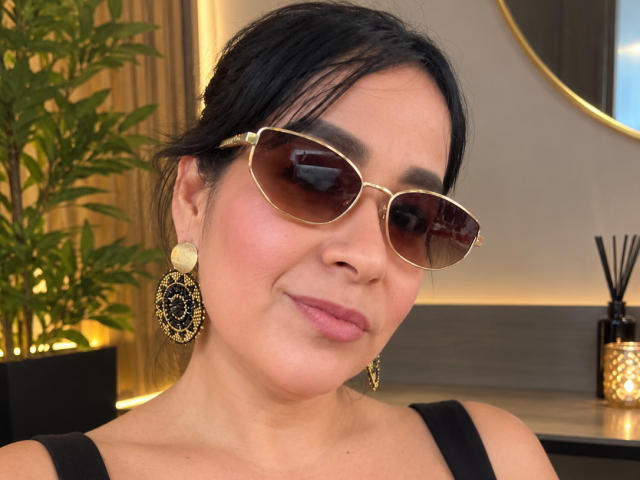 TamaraFoxy has black hair, brown sunglasses, gold earrings with black patterns, and pink lipstick. She's smiling slightly with a black top. Her ass is not visible.