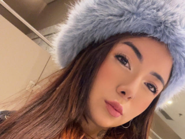 AriLynn with long brown hair, wearing a fluffy blue hat, has full pink lips and arched eyebrows. Her hoop earrings are visible. She's looking slightly to the side.