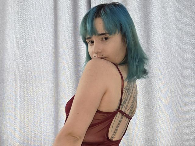 TeraHammar with teal blue hair, looking over her shoulder, wearing a sheer red top, showing a tattoo on her back, with her tits slightly visible.