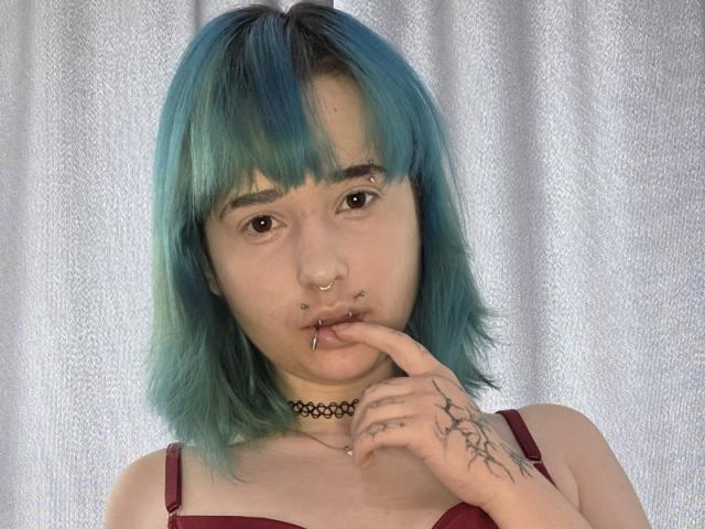 TeraHammar with teal-blue hair, nose ring, lip ring, finger in mouth, choker, tattoo on hand, wearing red bra, looking at camera.