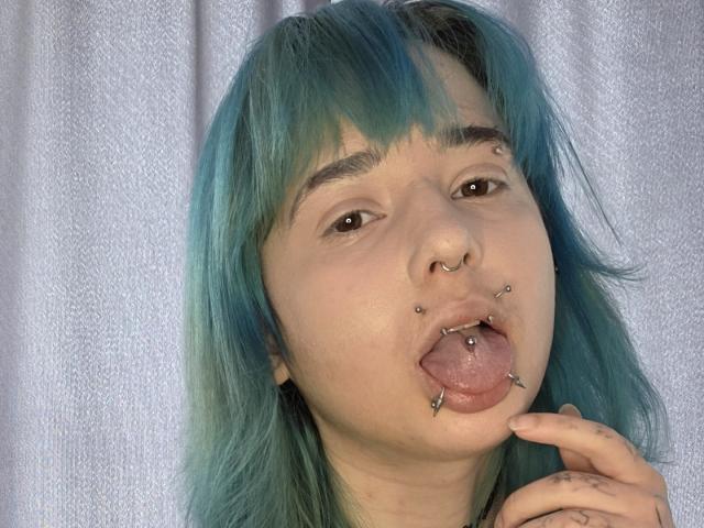 TeraHammar with teal hair, nose ring, and tongue piercings, sticking out her tongue, hand near mouth, looking at camera.