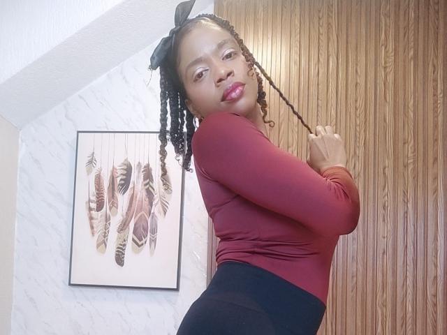 SarahiPusey with braided hair, red long-sleeve top, black high-waisted pants, holding a braid, looking over shoulder, pink lipstick, wooden wall, white and marble wall, framed art.