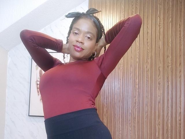SarahiPusey with dark skin, red long-sleeve top, black skirt, hands behind head, black hair in ponytail, red lipstick, smiling, medium tits, wooden wall, marble wall.