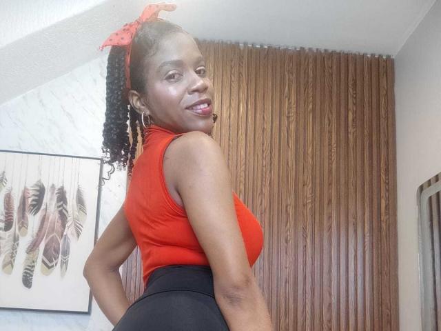 SarahiPusey has dark skin, curly black hair with a red bow, wearing a red sleeveless top and black high-waisted pants, showing her ass and smiling.