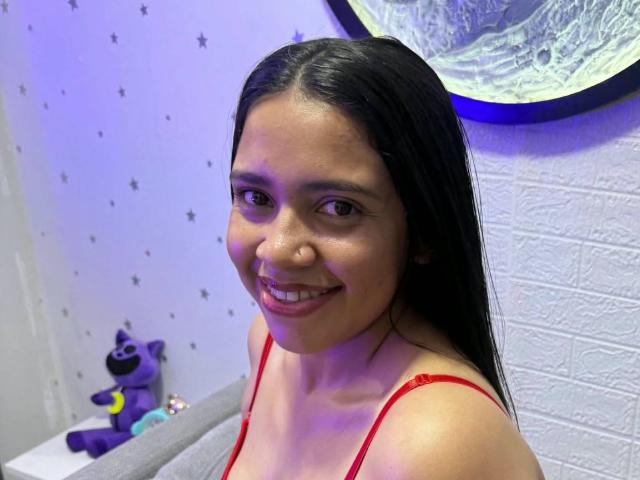 VickyRousse with long black hair, smiling, wearing a red spaghetti strap top, showing small tits, cute, medium brown skin, and a cute nose, in a brightly lit room.
