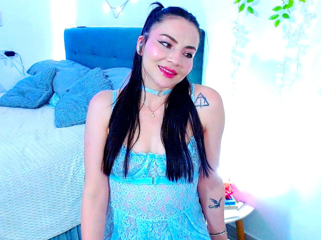 EmelySofia in a blue lace babydoll, smiling with pink lipstick, black hair in pigtails, small tits, tattoo on right shoulder, necklace, standing in front of a blue bed.