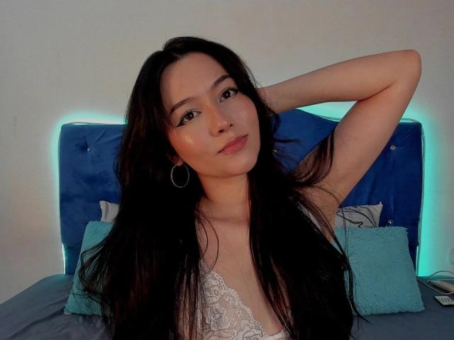 DeisyWest with long black hair, wearing a white lace top, has her arm raised, showing off her tits, and looking cute with a slight smile, sitting on a blue bed.