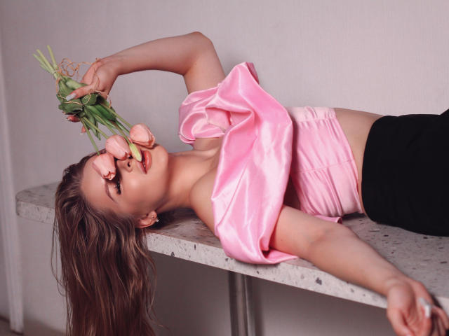 SnezhanaMeir laying on a table, wearing a pink satin top, holding pink roses to her nose, black pants, long brown hair, soft pink wall in background.