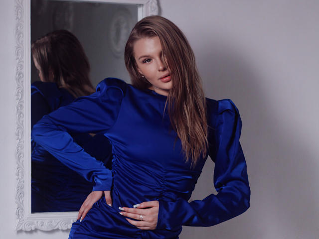 SnezhanaMeir in a shiny blue dress, hand on hip, long brown hair, looking at mirror, reflection shows back of dress, ring on finger, confident, sexy, elegant.