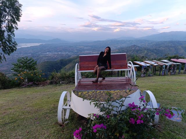 PauletteLoveX avec ses fesses sur un banc en bois, portant un haut noir et un short, assise sur une colline herbeuse, des fleurs violettes devant, une structure métallique blanche, un ciel de coucher de soleil, des montagnes en arrière-plan..