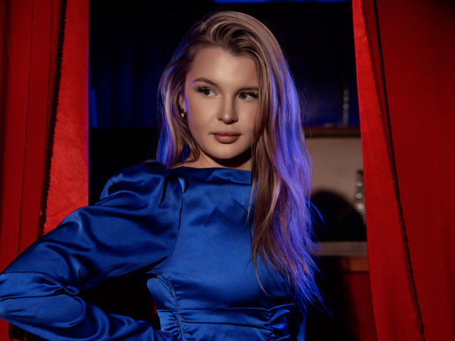 SnezhanaMeir with long blonde hair, wearing a shiny blue satin dress, standing in front of red curtains, looking directly at the camera with a slight smile.