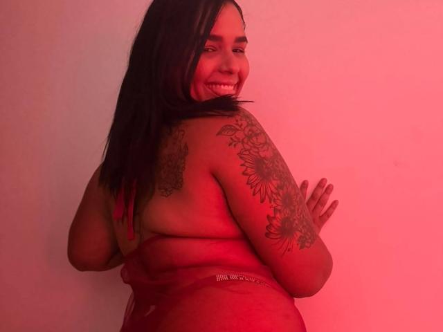 IvoneKing, topless, red-tinted, smiling, looking back, tattooed arm, black hair, medium tits, curvy ass, hands against wall, confident, sexy, soft lighting.