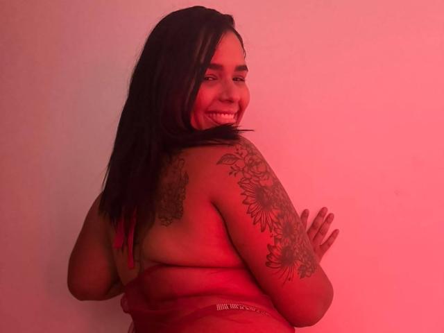 IvoneKing, con lunghi capelli neri, sorride, è senza top, mostrando tatuaggi sul braccio destro e sulla spalla, illuminazione rossa, e il suo sedere è parzialmente visibile..