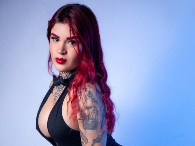 HalleyCloud tiene el cabello con puntas rojas, tatuajes en el brazo y lleva un vestido negro, de corte profundo, con un lazo, que muestra sus grandes senos y lápiz labial rojo..