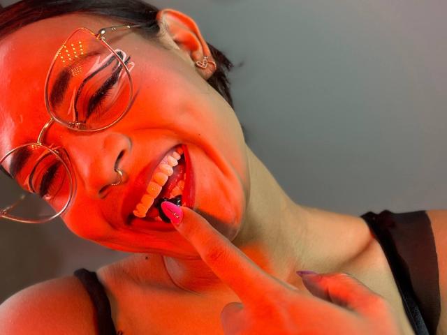 LilithRussoo with red lighting, smiling, biting pink nail, wearing glasses, nose ring, and black strap dress, showing white teeth, ear piercing, and pink nail polish.